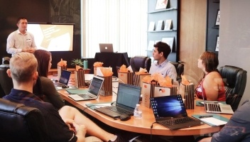 Several businesspeople sitting around a conference table with laptops open in front of each, listening to the meeting leader.