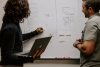 Two individuals make user experience (UX) research notes using a laptop and whiteboard.