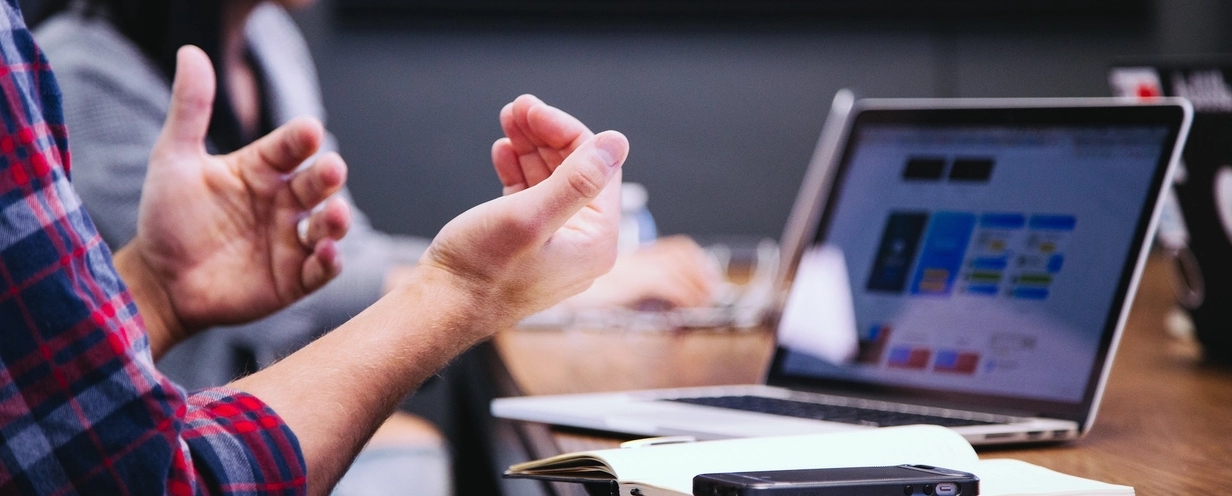 Photo of two people sitting near a laptop. One person is gesturing with their hands. 