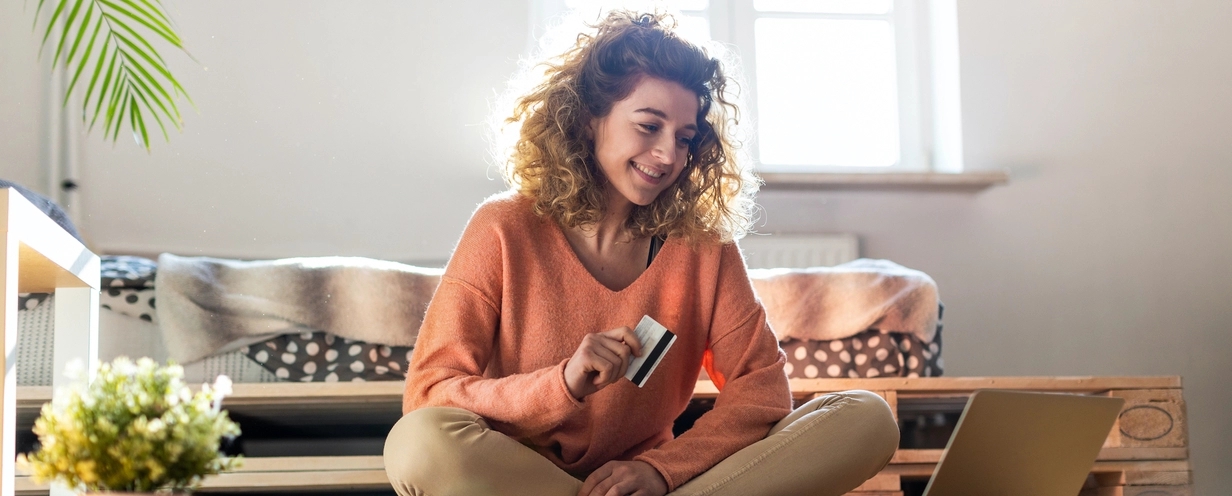 Photo of a woman sitting on the floor of a well lit room holding her credit card while looking at her laptop
