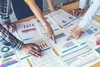 Team of 3 people pointing to charts and graphs on sheets of paper on a conference table