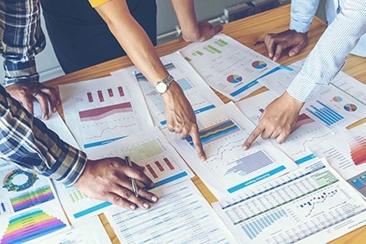 Team of 3 people pointing to charts and graphs on sheets of paper on a conference table